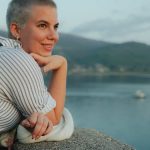 A smiling woman with short hair leans on a wall, enjoying a scenic coastal view.